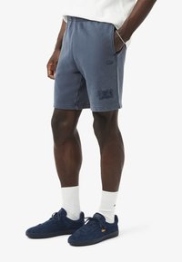 Shorts in felpa blu navy con vita elastica, tasche laterali e una toppa con il logo Lacoste. Abbinati a sneakers in suede blu navy e calzini bianchi.