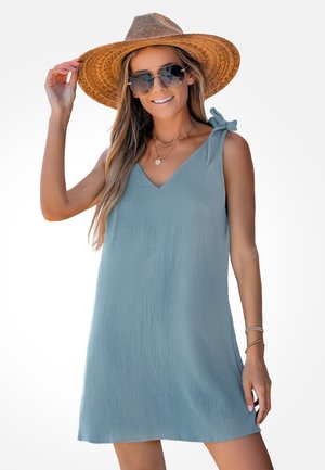 Frau trägt ein hellblaues ärmelloses Kleid mit Schulterbindung, großen Strohhut, Sonnenbrille und geschichtete Halsketten, lächelt vor weißem Hintergrund.