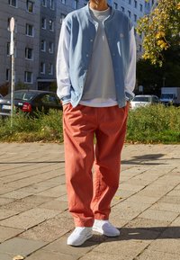 Jasnoniebieska, zapinana kurtka w stylu varsity, szary T-shirt, owerzoowe koralowe spodnie i białe sneakersy, na tle miejskiego krajobrazu z zielenią.