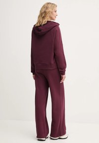 Burgundská mikina s kapucňou a voľným strihom, dlhé rukávy, doplnená zodpovedajúcimi širokými nohavicami. Hladká zmes bavlny.