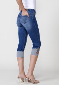 Jean capri en denim bleu foncé avec un motif floral brodé sur les ourlets roulés et une silhouette ajustée, comprenant un style à cinq poches.