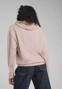 Sudadera rosa con capucha, con hombros caídos y puños de canalé, combinada con pantalones de mezclilla oscuros que presentan costuras visibles y bolsillos.