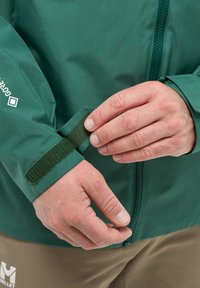 Veste imperméable verte avec une texture lisse, poignet ajustable en Velcro et fermeture éclair à l'avant. Présente le logo de la marque sur la manche.