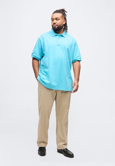 Άνδρας με dreadlocks που φοράει φωτεινό γαλάζιο πόλο πουκάμισο, μπεζ παντελόνι και μαύρα μοκασίνια, στέκεται με το ένα χέρι στην τσέπη ενάντια σε λευκό φόντο.