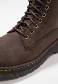 Bota de cuero marrón con ojales metálicos, suela negra gruesa, costuras amarillas y superficie texturizada. Los cordones están enhebrados a través de los ojales.