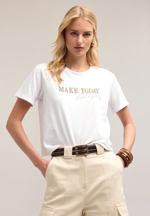 Witte t-shirt met korte mouwen featuring een metallic gouden grafiek met de tekst "MAAK VANDAAG Geweldig." Gecombineerd met beige high-waisted broeken en een bruine riem.