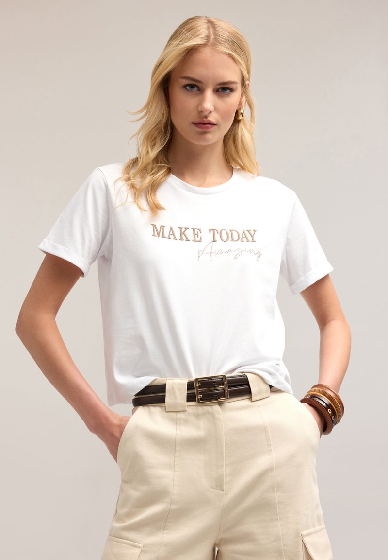 Vit kortärmad t-shirt med en metallisk guldgrafik som säger "GÖR IDAG Fantastisk." I kombination med beige högmidjade byxor och ett brunt bälte.