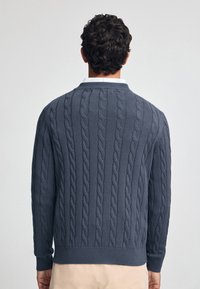 Marinblå kabelstickad tröja med ribbad nederkant och ärmslut, med en klassisk krage och texturerat mönster över hela ryggen.