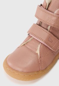 Zapato de piel rosa con tiras de velcro y suela de goma marrón claro. Presenta costuras visibles y un forro interior suave. Diseño de punta redondeada.