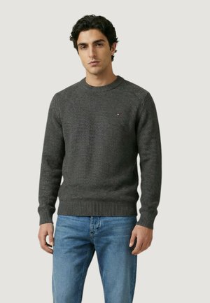 Uomo che indossa un maglione girocollo texturizzato grigio scuro e jeans azzurri, in piedi contro uno sfondo chiaro e semplice.