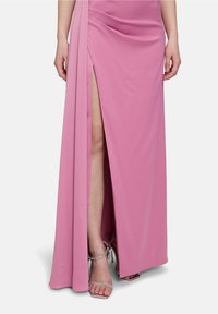 Roze jurk tot op de grond met een zij-split en gedrapeerd stofdetail. Gecombineerd met zilveren enkelsandalen met bandjes. Soepele textuur, elegante uitstraling.