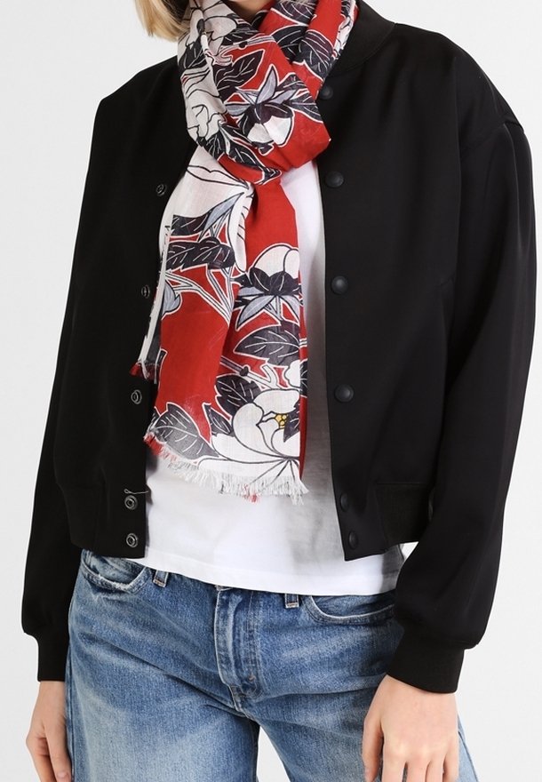 Personne portant une veste noire boutonnée, une chemise blanche, un jean bleu et une écharpe rouge à motif floral autour du cou.