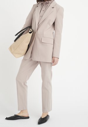 Person trägt beigen Anzug und Bluse und hält große beige geflochtene Handtasche mit schwarzen Griffen, steht in schwarzen spitzen flachen Schuhen.