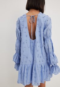 Svetlo modra obleka z globokim izrezom na hrbtu in s povezovanjem. Ima cvetlični vez, dolge bujne rokave in nabrano obrobo.