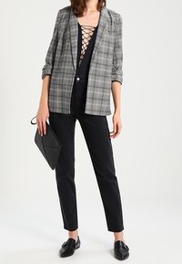 Grau kariertes Sakko über einem schwarzen Oberteil mit Schnür-Ausschnitt, kombiniert mit schwarzen Hosen und flachen schwarzen Schuhen, hält eine schwarze Clutch.