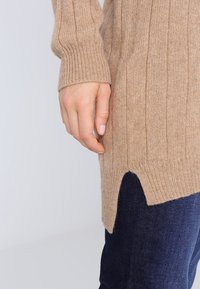 Main reposant contre la cuisse, portant un pull côtelé beige avec une fente latérale et un jean bleu foncé, sur fond clair.