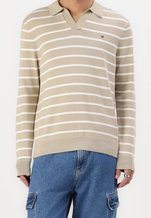 Chemise en maille à manches longues beige et blanche rayée avec col, portée avec un jean cargo bleu, présentant un petit logo sur le côté gauche de la poitrine.