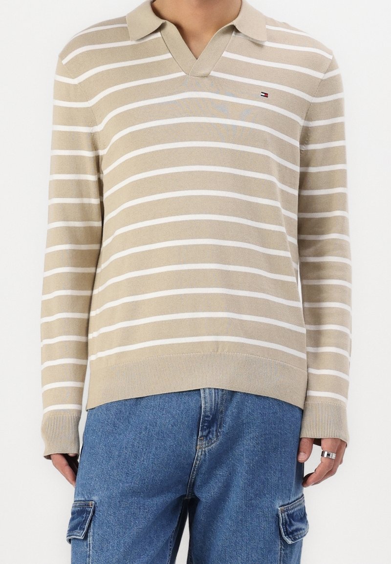Chemise en maille à manches longues beige et blanche rayée avec col, portée avec un jean cargo bleu, présentant un petit logo sur le côté gauche de la poitrine.