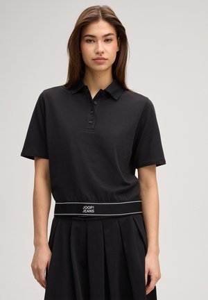 Mladá žena s rovnými hnedými vlasmi nosí čierne polo tričko s krátkym rukávom a plisovanú sukňu s opaskom JOOP! Jeans v páse.