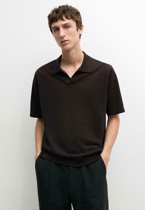 Jeune homme aux cheveux courts et bouclés, portant une chemise à manches courtes marron foncé avec col et un pantalon sombre, debout les mains dans les poches.