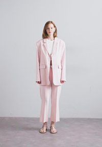 Blazer ajusté rose clair avec un pantalon droit assorti, associé à un top blanc et des sandales à enfiler, tissu lisse, design minimaliste.