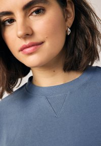 Modrá bavlnená mikina s okrúhlym výstrihom, rebrovaným lemom a detailom textúrovaného šitia. Modelka má na sebe strieborné visiace náušnice.