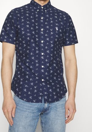 Man draagt een marineblauw kort mouw overhemd met kleine witte nautische en fiets patronen, gecombineerd met lichtblauwe spijkerbroek.