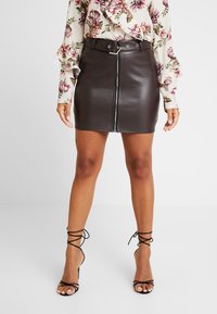 Brun fusk läder mini kjol med frontdragkedja och spännbelt detaljer, parat med svarta snörade klackar och en blommig blus.