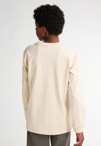 Tricou cu mâneci lungi din bumbac bej deschis, cu guler rotund și mansete elastice. Are un design simplu, fără modele sau grafice vizibile.