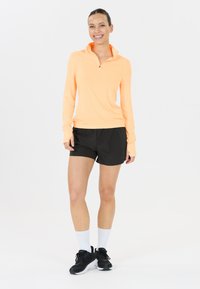 Leichtes, langärmeliges, mit Reißverschluss versehenes Oberteil in Neonorange, kombiniert mit schwarzen Shorts, weißen Socken und schwarzen Sportschuhen. Glatte Textur.