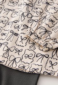 Cream fabric with a textured surface features a black bow and scalloped pattern, complemented by a dark ribbed hem.