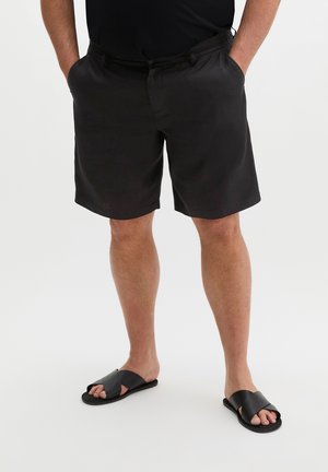 Man draagt zwarte casual shorts en zwarte instapsandalen, staat met handen in de zakken tegen een effen witte achtergrond.