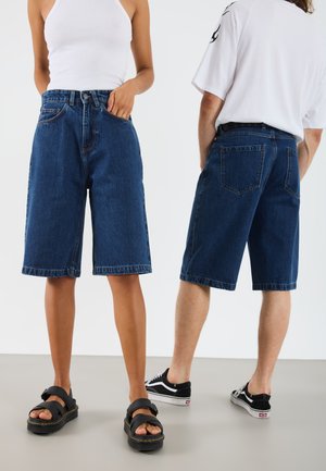 Denim shorts in dark wash with raw hem, loose fit, five pockets, and visible stitching details, paired with sandals and casual shoes.