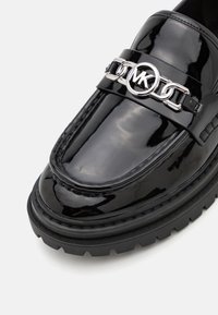 Mocassin en cuir verni noir avec un logo MK rond en argent et un détail en chaîne, présentant une semelle en caoutchouc épaisse et des surpiqûres en relief.