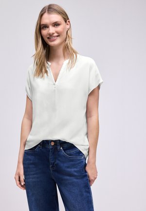 Smiling woman with long blonde hair wears a loose white short-sleeve blouse and dark blue jeans against a plain light background.