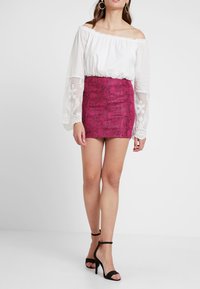 Witte off-shoulder blouse met lange, doorschijnende mouwen en bloemenborduursel, gecombineerd met een strakke roze minirok met slangendessin en zwarte hakken.