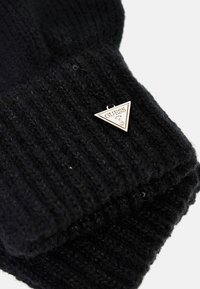 Juodas megztas kepurė su tekstūruotu paviršiumi ir trikampiu sidabro metalo logotipo ženkleliu su užrašu "GUESS". Medžiaga atrodo minkšta ir šilta.