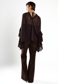 Blusa de chiffon negra fluida con mangas con volantes, escote en V en la espalda y detalle atado. Combinada con pantalones de campana marrones. Diseño minimalista.