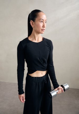 Black long-sleeve cropped top with a twist detail, paired with high-waisted black pants. Holding a silver dumbbell with a matte finish.