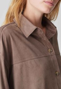 Brown suede shirt with a collared neckline, front button closure, and visible stitching details. Soft texture and relaxed fit.