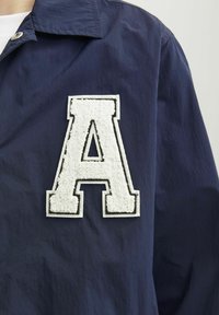 Mornarska jakna z teksturirano belo aplikacijo črke "A" obrobljeno z črno. Narejena iz gladkega blaga z ovratnikom in gumbnim zapiranjem.