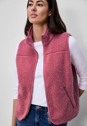 Vrouw die een mouwloze roze sherpa fleece bodywarmer draagt over een wit shirt met lange mouwen en blauwe spijkerbroek, staand tegen een effen achtergrond.