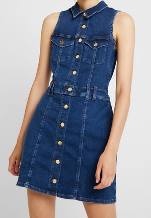 Bezrekawowa ciemnoniebieska sukienka z denimu z złotymi guzikami z przodu, kieszeniami na piersi oraz kołnierzykiem, noszona przez stojącą osobę.