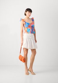 Haut à imprimé floral coloré avec des bretelles volantes, associé à une jupe plissée blanche. Les accessoires comprennent un sac bandoulière orange et des talons beige à lanières.
