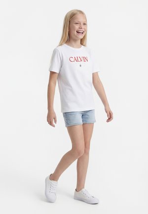 Lachend meisje loopt, draagt een wit Calvin T-shirt, lichte denimshorts en witte sneakers tegen een effen witte achtergrond.