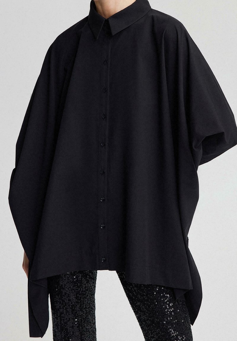 Chemise noire ample à boutons avec un ourlet étendu et asymétrique portée sur un pantalon noir à sequins.