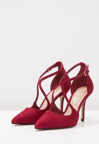 Ett par röda mocka högklackade skor med spetsig tå, T-remsa och ankelsnärning på vit yta mot ljus bakgrund.