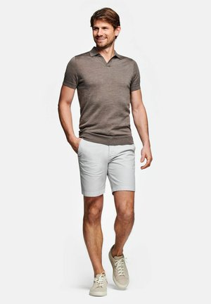 Man loopt, draagt een bruin kort mouw gebreid polohemd, lichtgrijze korte broek en beige sneakers, glimlacht en kijkt opzij.