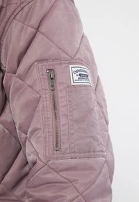 Purple quilted jacket with a zippered pocket on the left sleeve. Features a label patch with "Original" text and silver hardware accents.