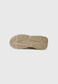 Tan sneaker sole with textured rubber, featuring a wave-like design and prominent brand logo. Smooth edges and multi-directional tread for grip.